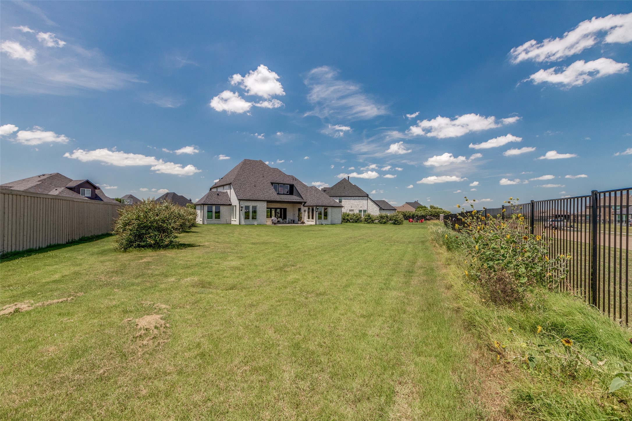 Large stone home beyond a broad grassy backyard, with a covered patio and fences on both sides.