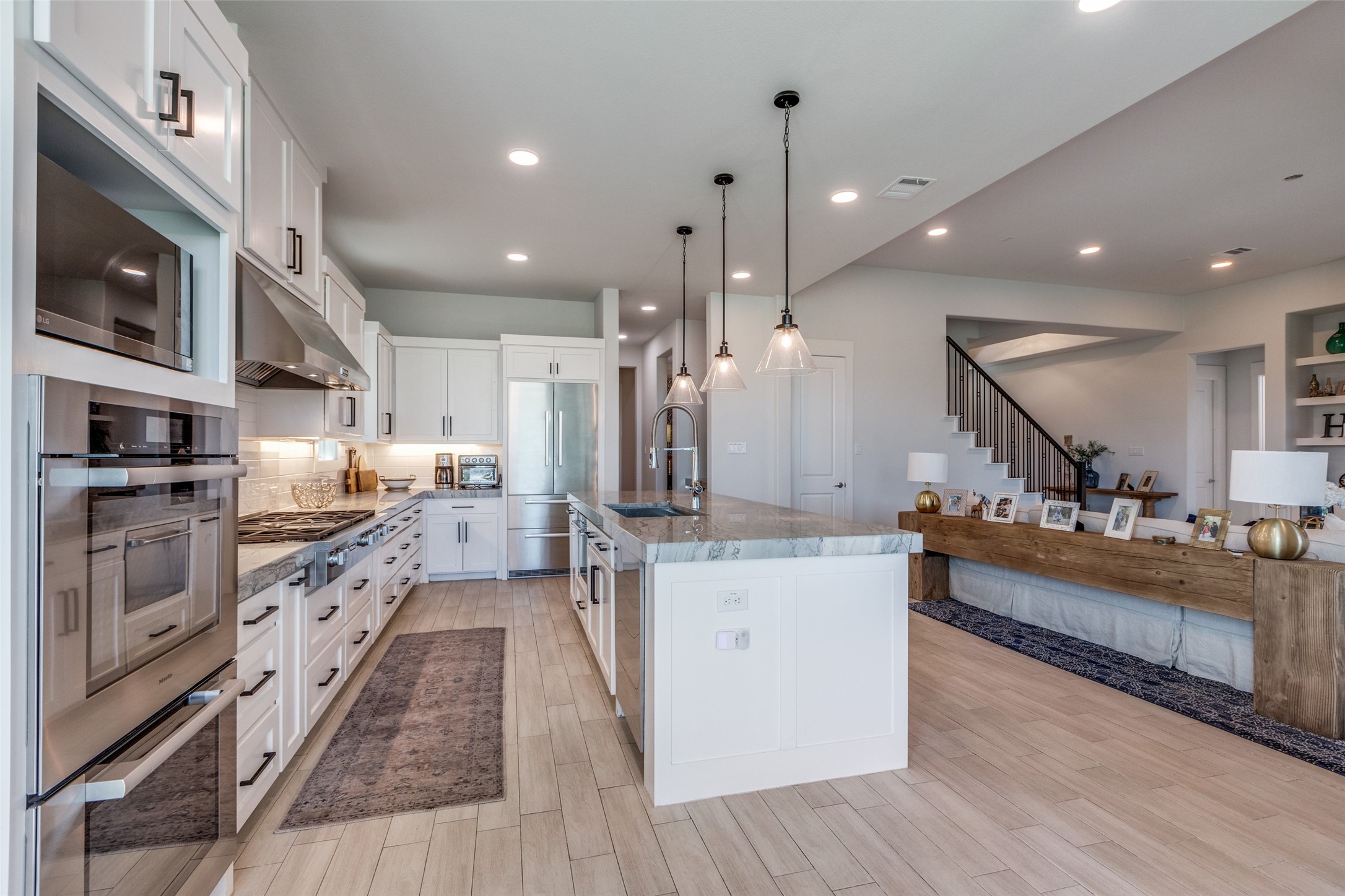 Open kitchen with a marble island, stainless steel appliances, and adjoining living area beneath a staircase.