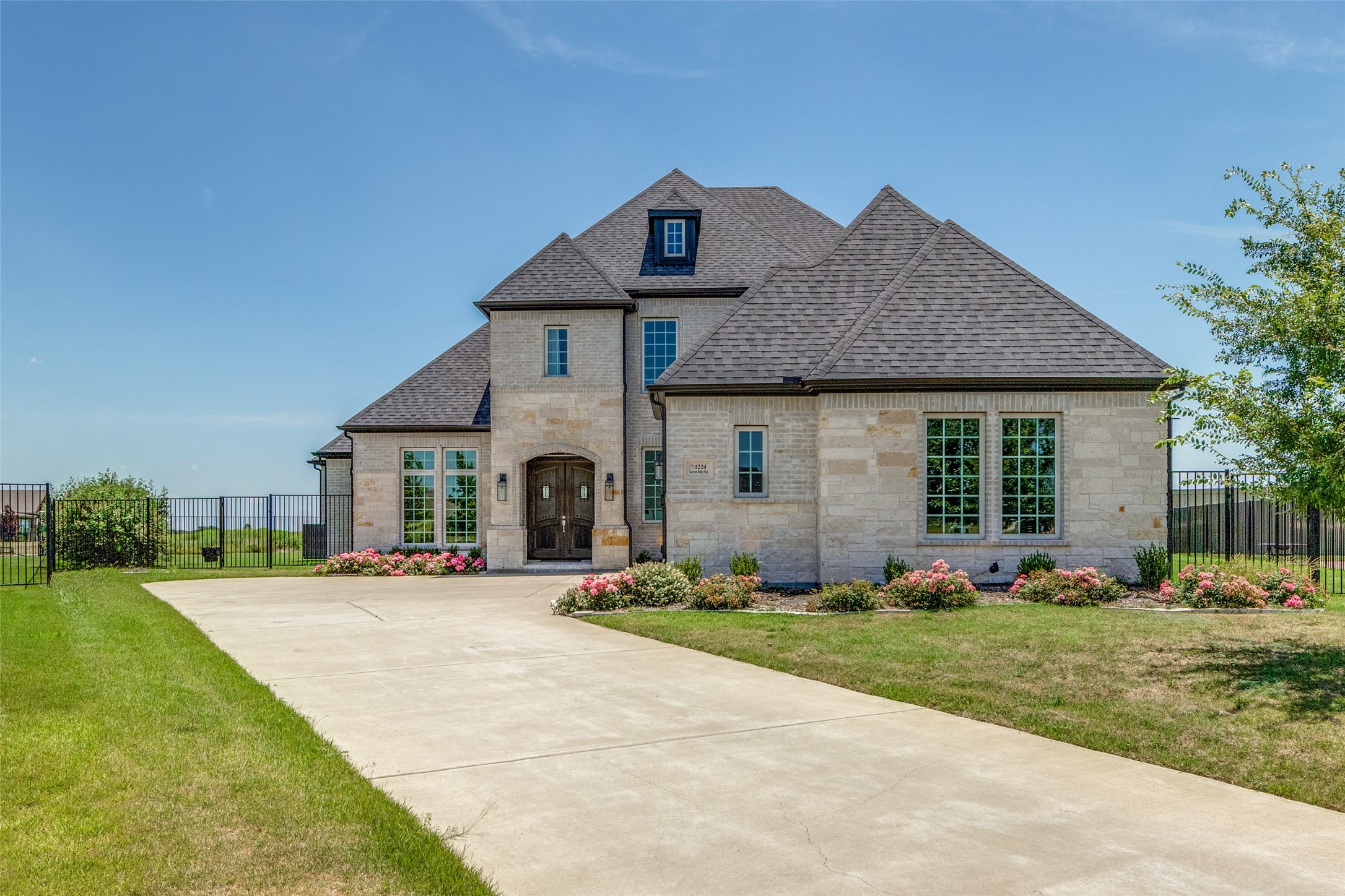 Stone house with multiple gables, arched entry, wide driveway, and pink flower beds.