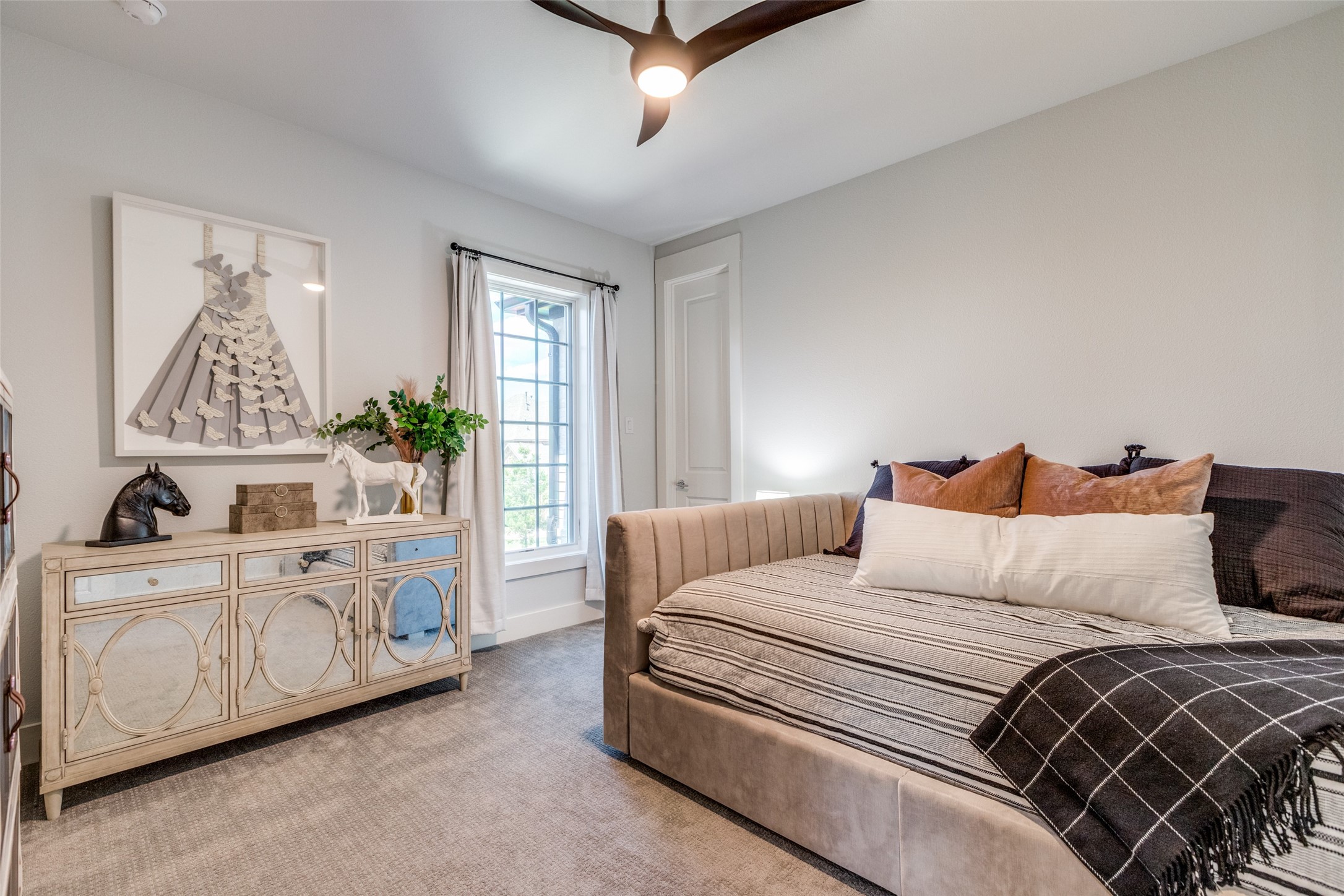 Furnished bedroom with an upholstered bed, rust-colored pillows, and a decorative dresser beside a tall window.