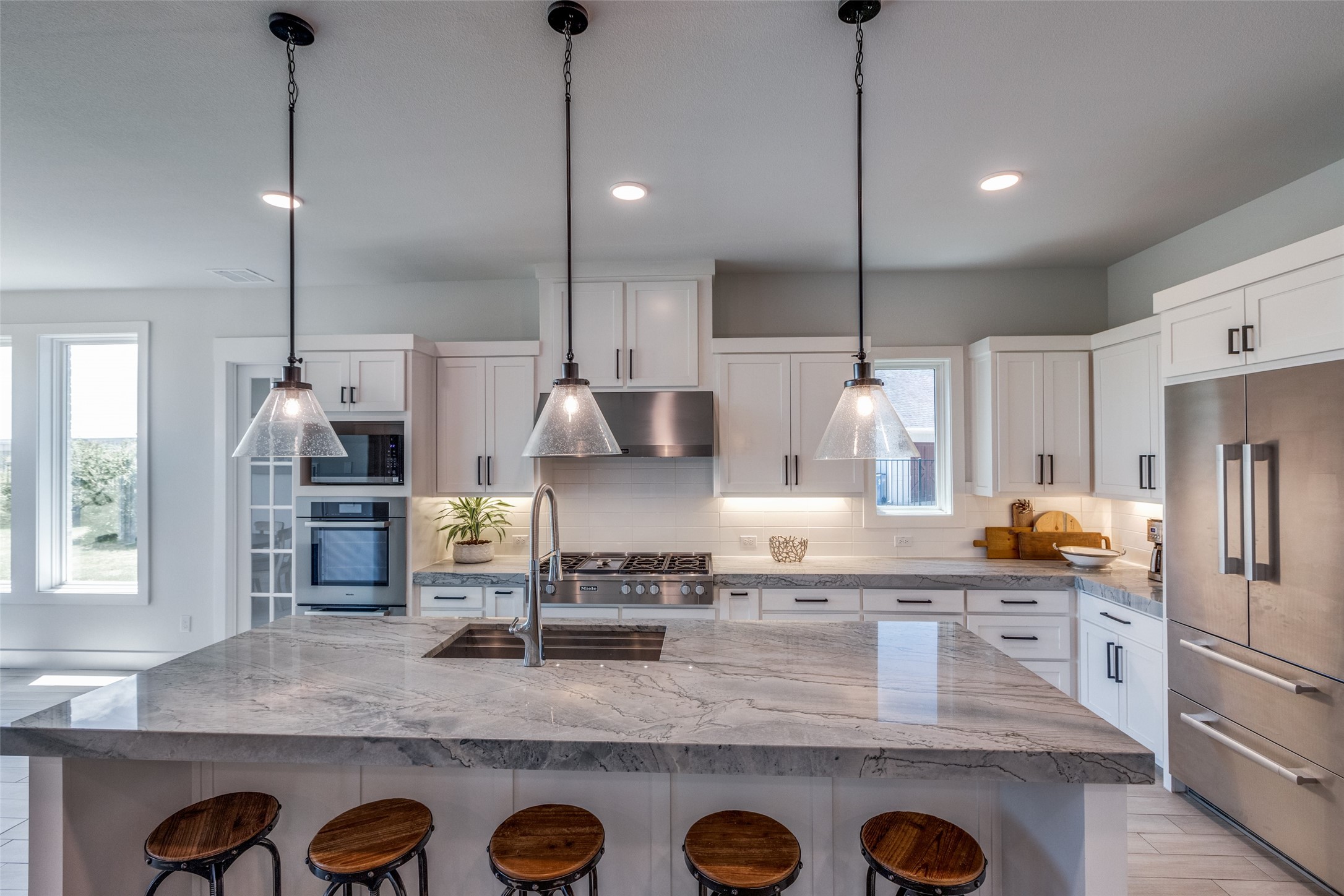 Modern kitchen with a large veined stone island, five wooden stools, and white cabinets.