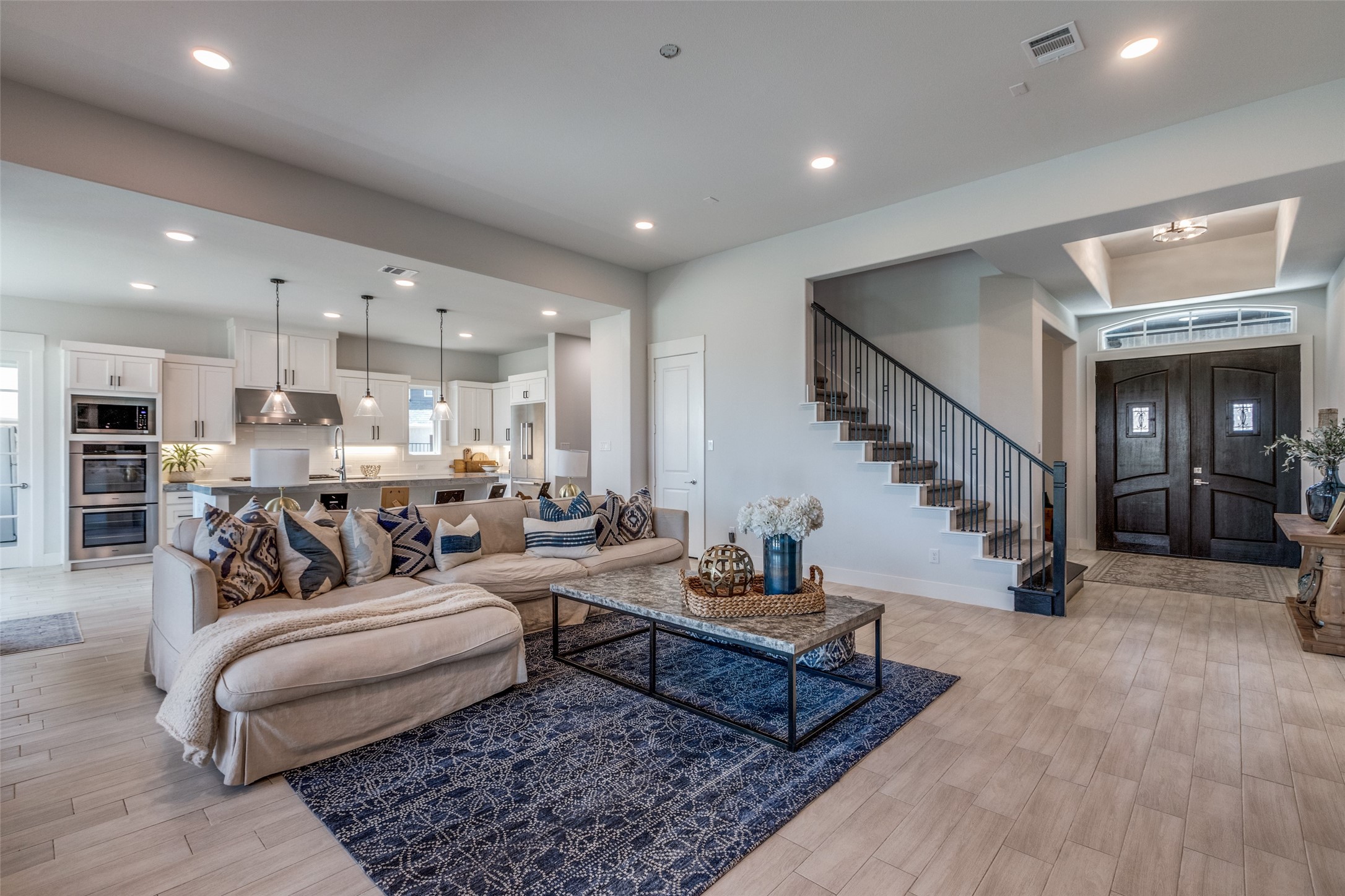 Open-plan living room with a light sectional, patterned navy rug, white kitchen, and staircase by the entry.