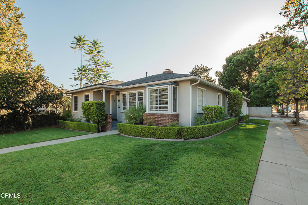 Single-story stucco home with a bay window, brick accents, clipped hedges, and a broad lawn.