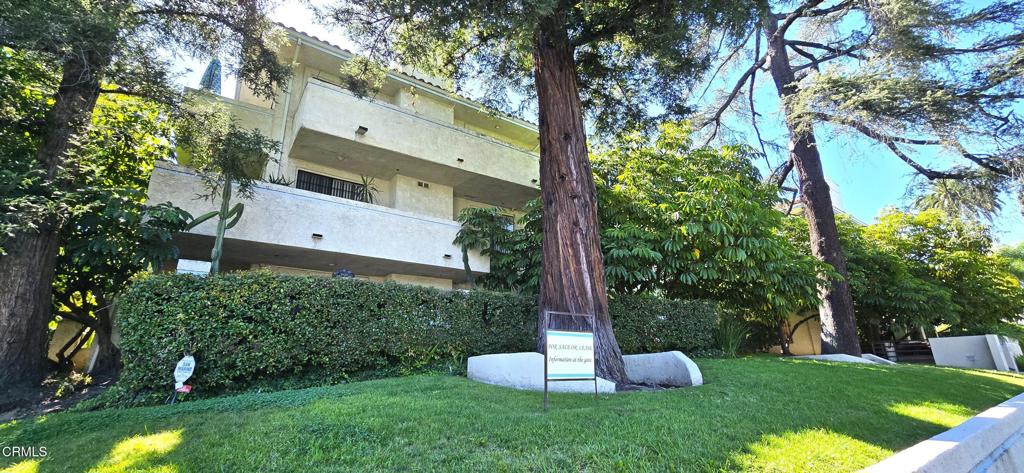 Residential building with balconies behind mature trees, hedges, and a front lawn.