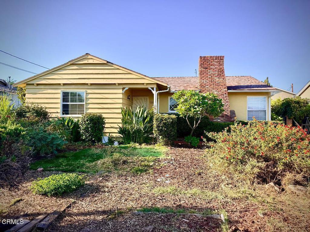 Single-story yellow house with horizontal siding, a brick chimney, and a leafy front yard.
