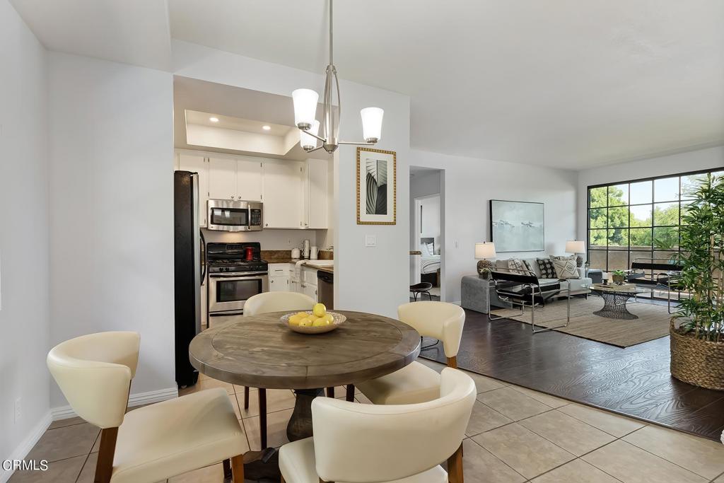 Open-plan dining area with a round table, white kitchen, and living room by large windows.