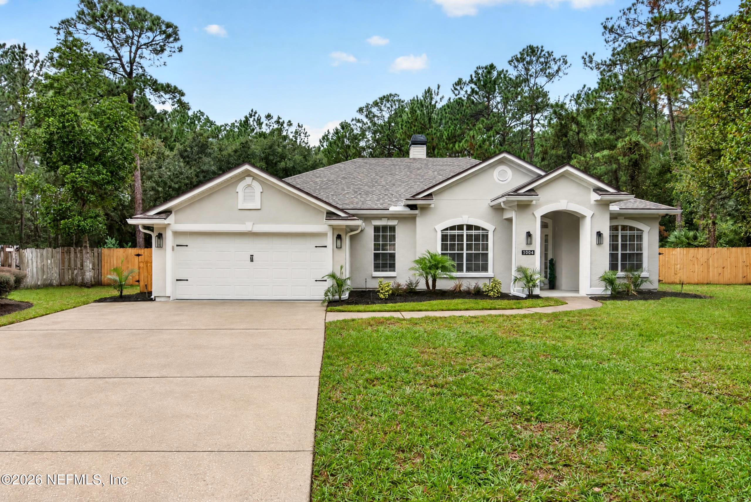Single-story home with a two-car garage, arched entry, and mature pine trees behind it.