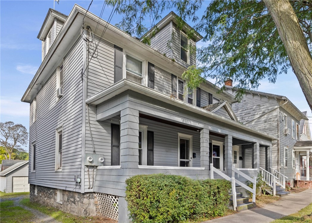 Gray multi-unit house with a broad covered porch, upper dormers, and a stone foundation.