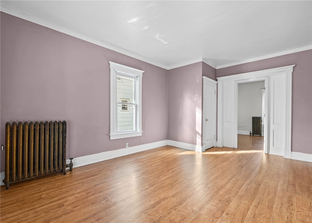 Vacant room with mauve walls, hardwood floors, a tall window, and a cast-iron radiator.