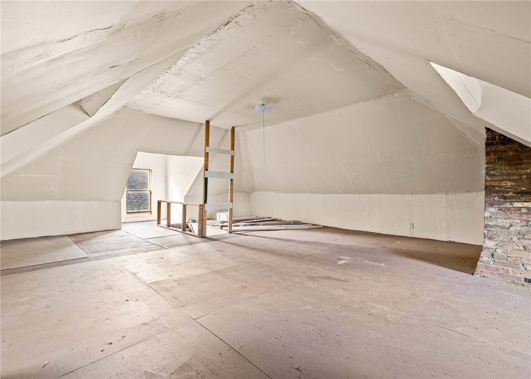 Unfinished attic with sloped white walls, plywood flooring, and exposed brick.