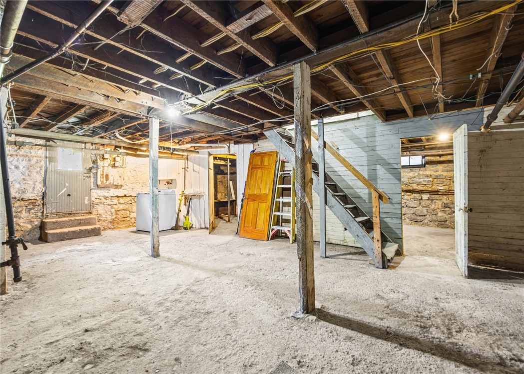 Unfinished basement with exposed ceiling joists, stone foundation walls, and a wooden staircase.