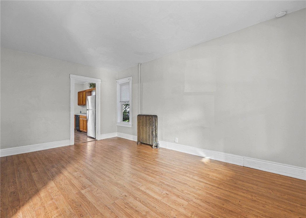 Empty room with hardwood floors, a vintage radiator, and a doorway to the kitchen.
