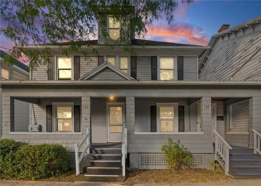 Gray clapboard two-story homes with lit windows and covered porches beneath a pink sunset sky.