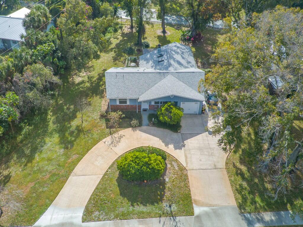 Aerial view of a single-story house with a gray roof and curved driveway amid mature trees.