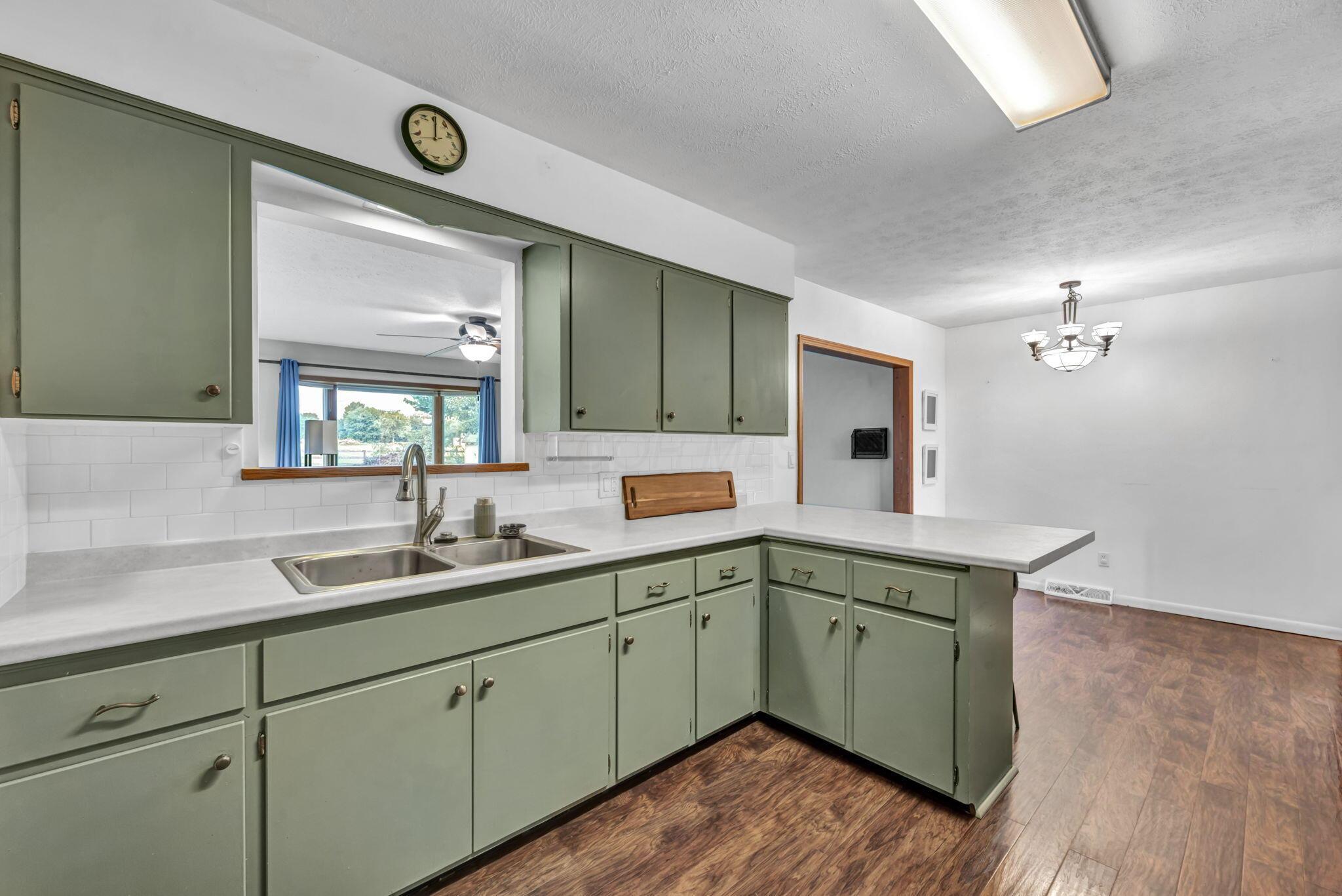 Kitchen with sage-green cabinets, a double-basin sink, and a wide pass-through to a window with blue curtains.
