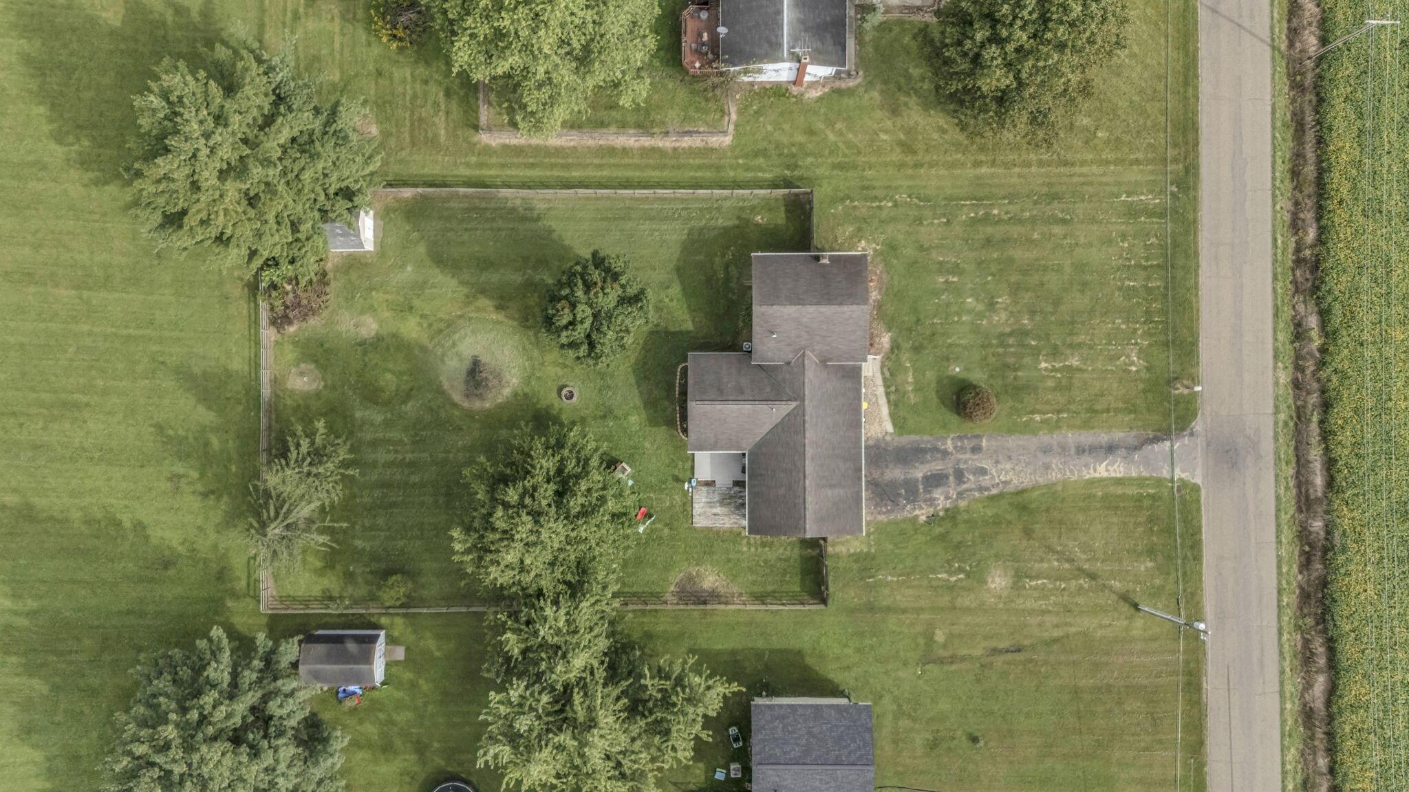 Aerial view of a house with fenced lawn, mature trees, and a roadside driveway.