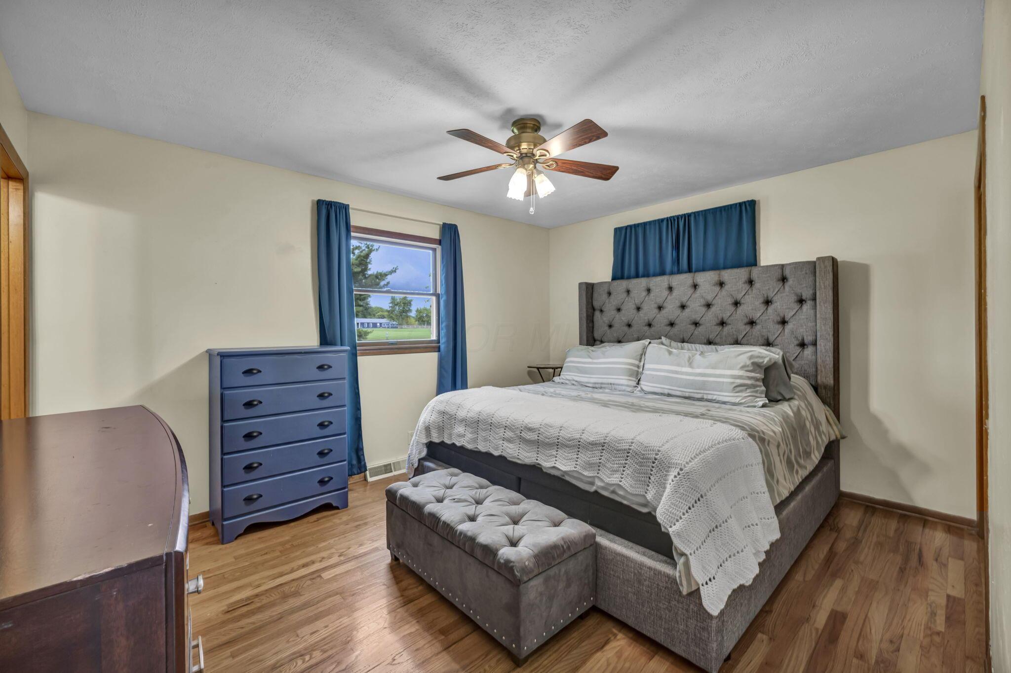 Bedroom with a gray tufted bed, upholstered bench, blue curtains, and hardwood floors.