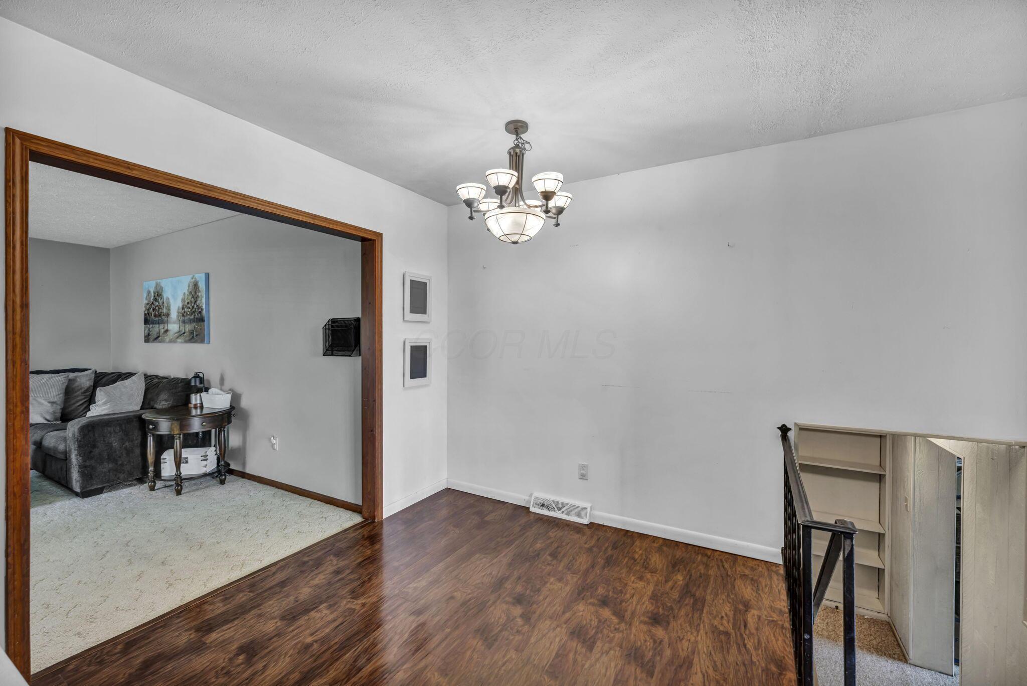 Dining area with dark wood flooring and chandelier, opening into a carpeted living room beside the stairs.