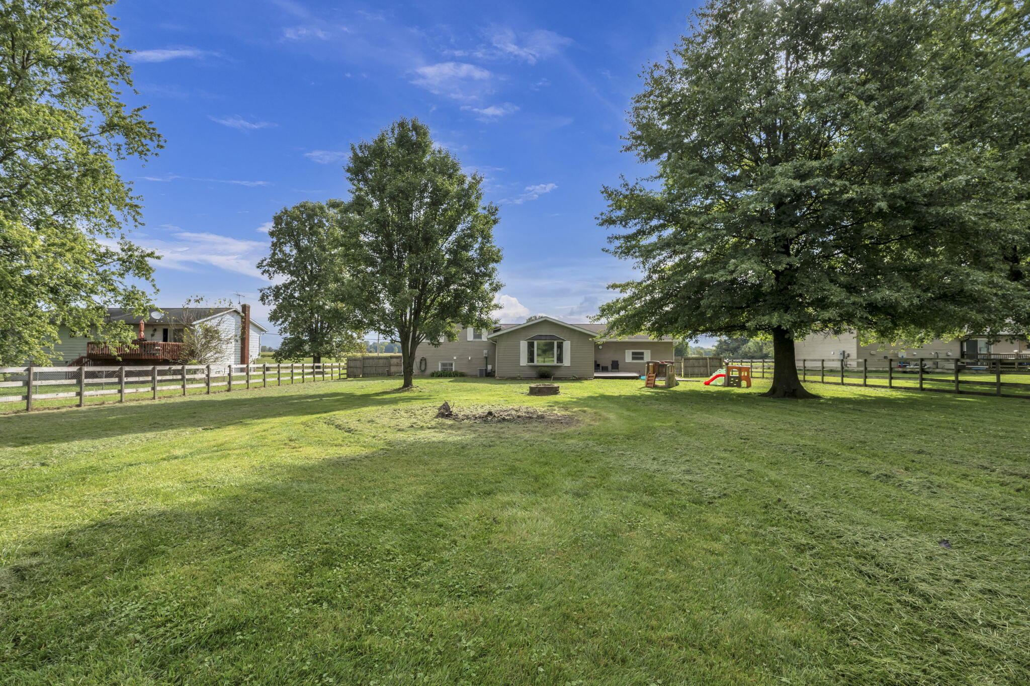 Backyard lawn and single-story home framed by mature trees, with a fenced yard, fire pit, and play set.