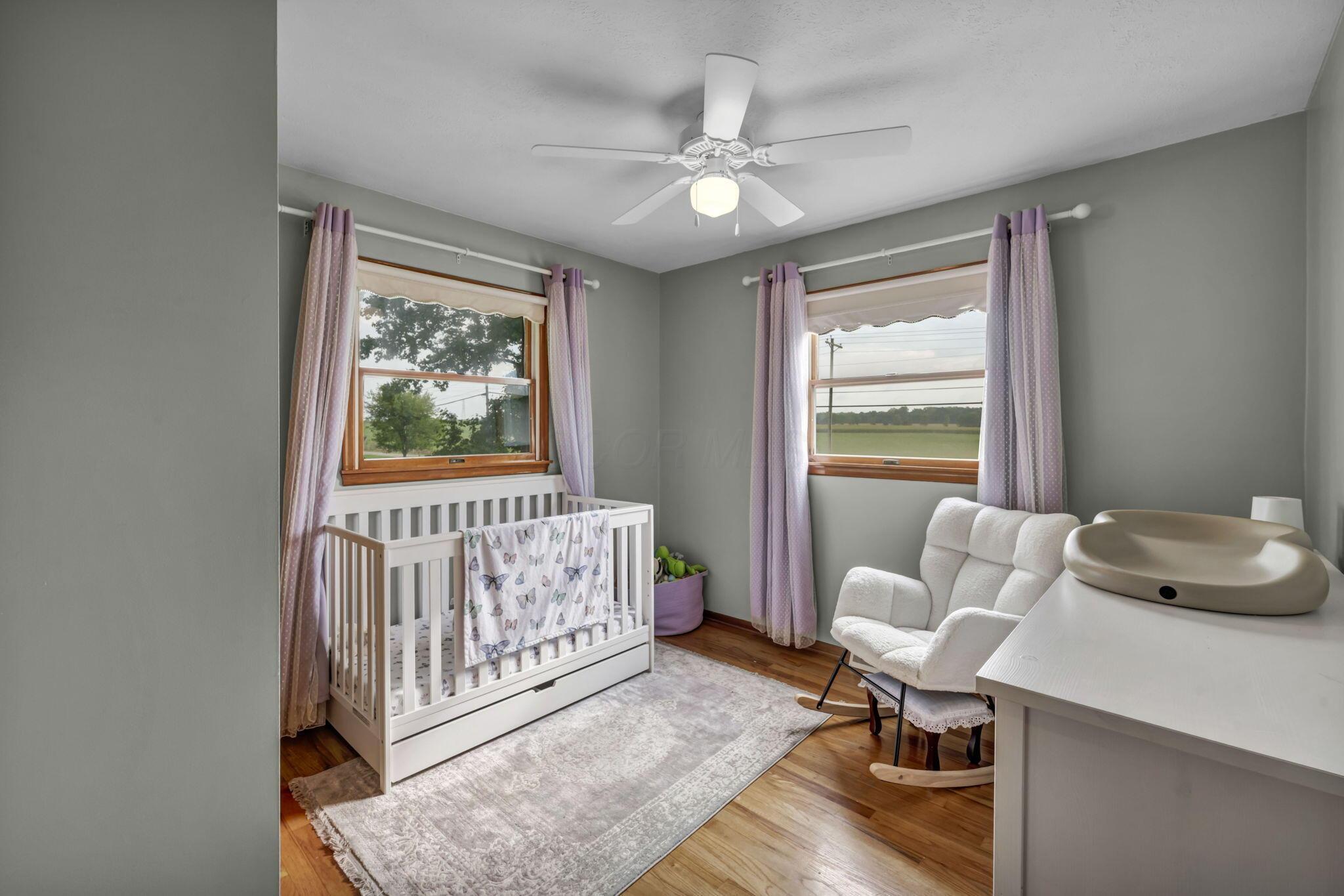 Nursery with a white crib, glider chair, and changing table beside windows overlooking open fields.