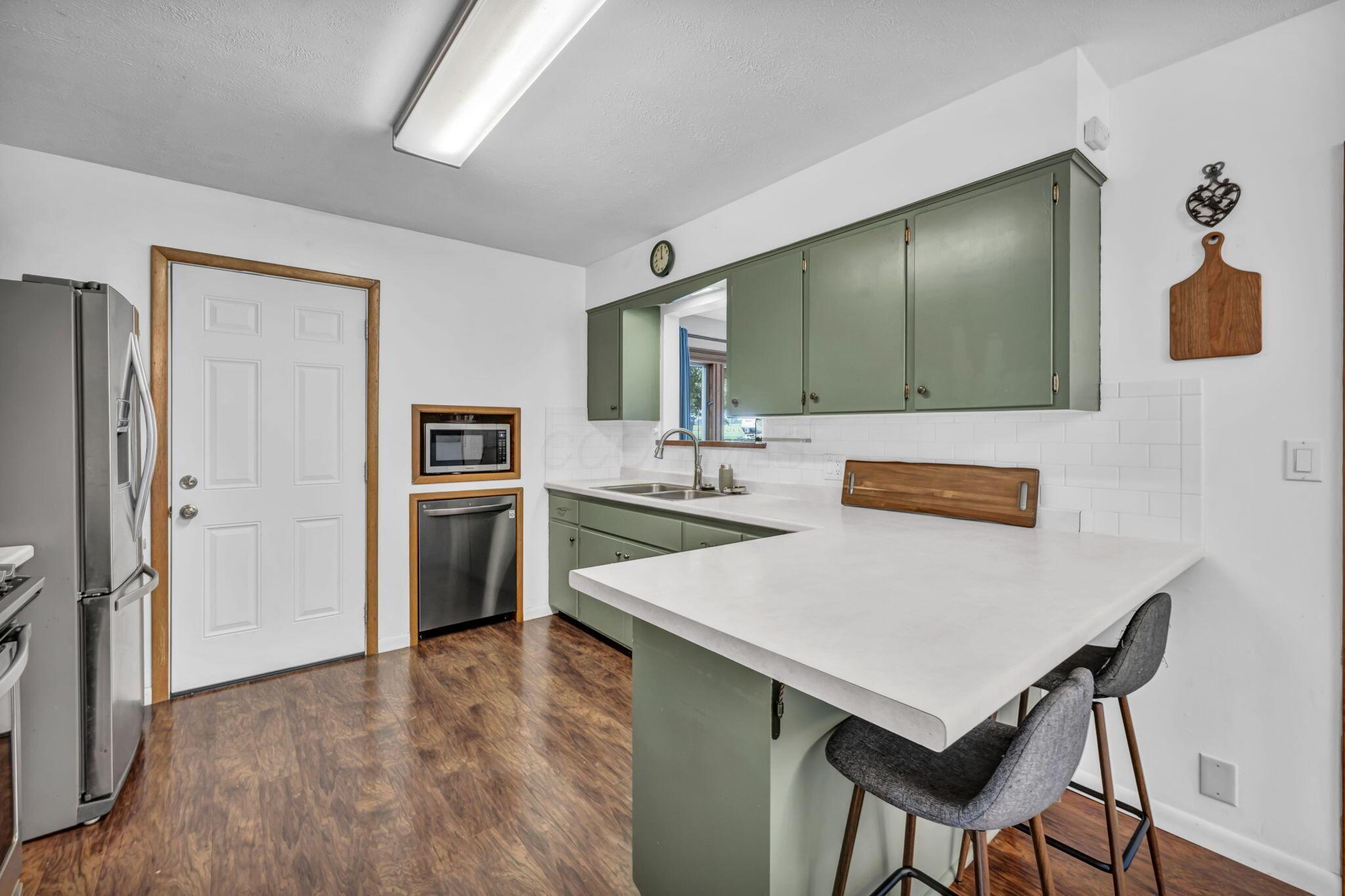 Kitchen with sage-green cabinets, white peninsula counter, stainless appliances, and wood-look flooring.