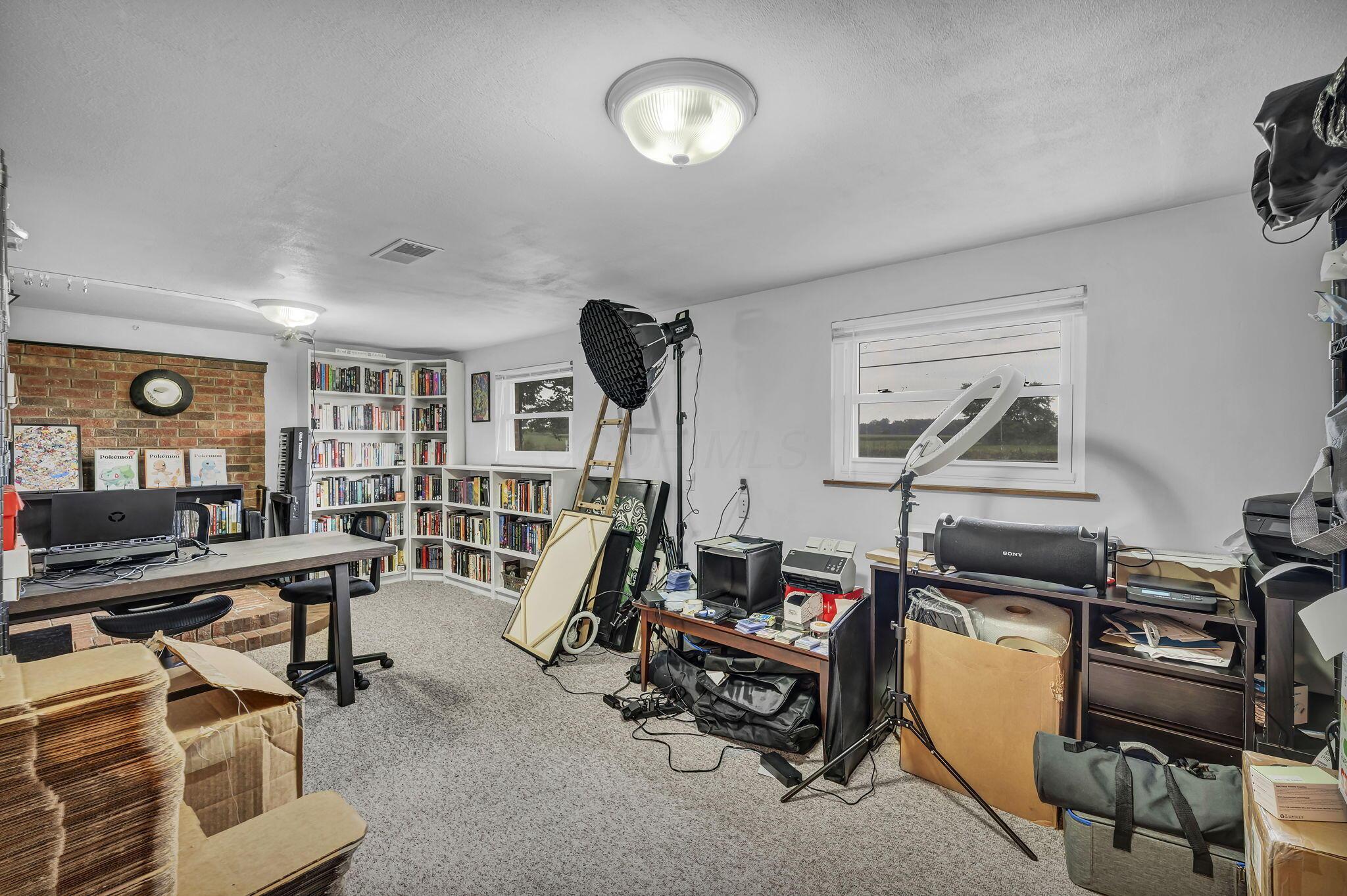 Home office with bookcases, a brick accent wall, and photography lights among boxes and equipment.