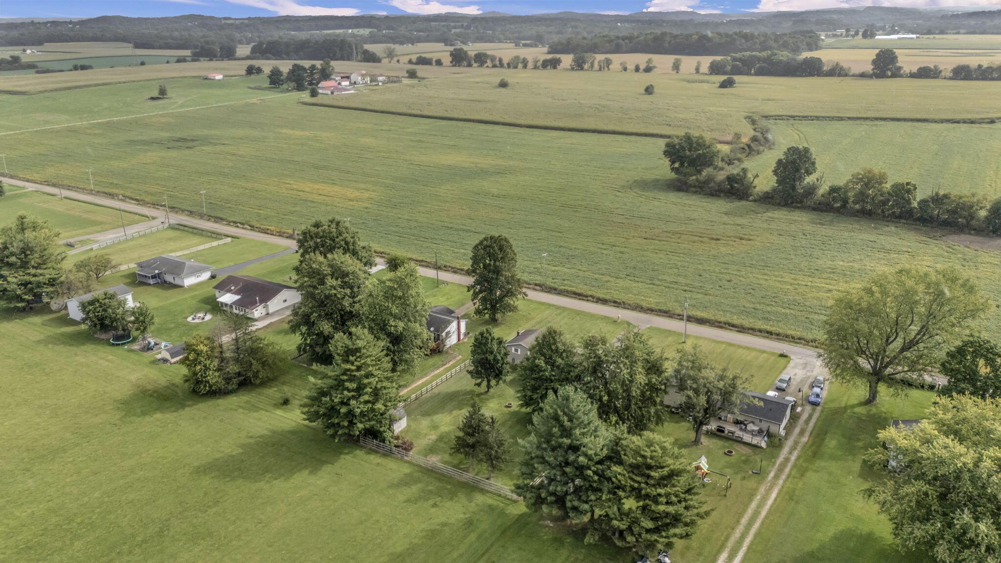 Aerial view of green farmland and scattered homes, with tree-lined roads and wooded hills beyond.