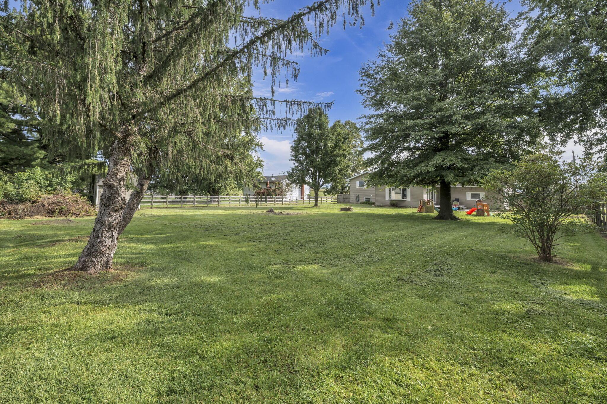 A spacious grassy yard with mature trees, a fenced boundary, and a one-story house.