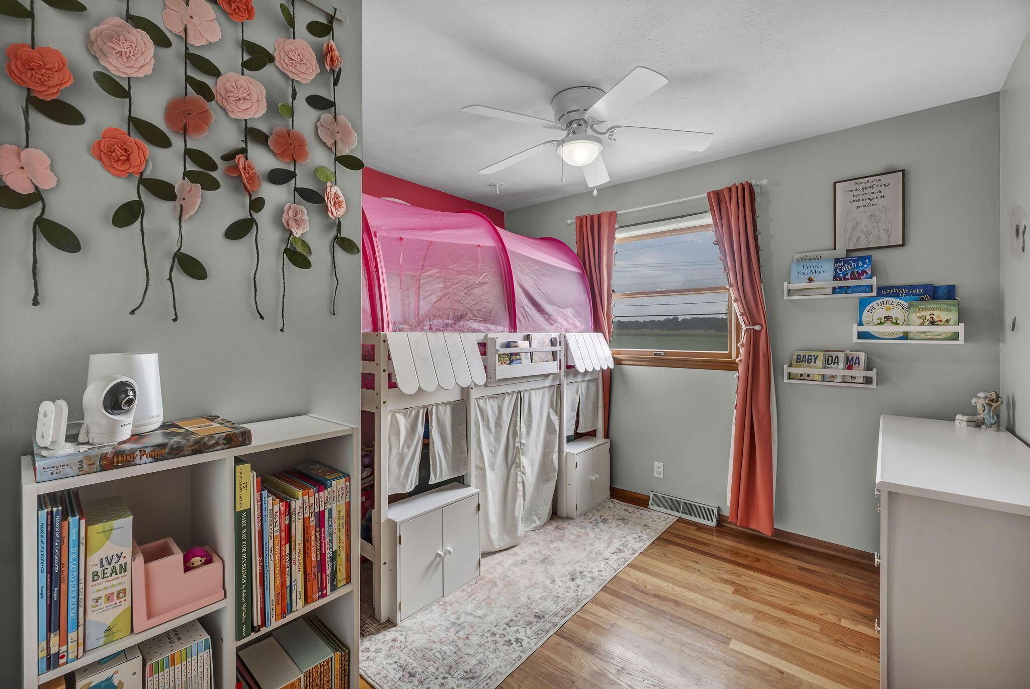 Child's bedroom with a pink canopy bunk bed, flower wall decorations, bookshelves, and a window overlooking fields.