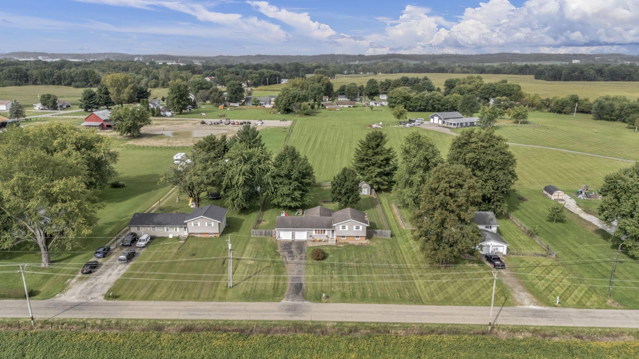 Aerial view of a rural home with a long driveway, broad lawns, trees, and open fields.