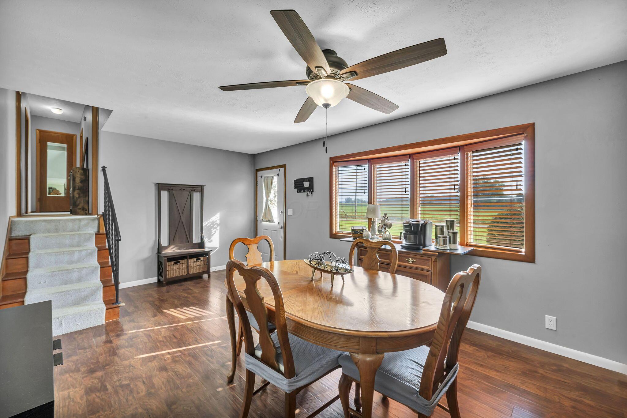 Dining room with a round wooden table, large windows overlooking fields, and a carpeted staircase.