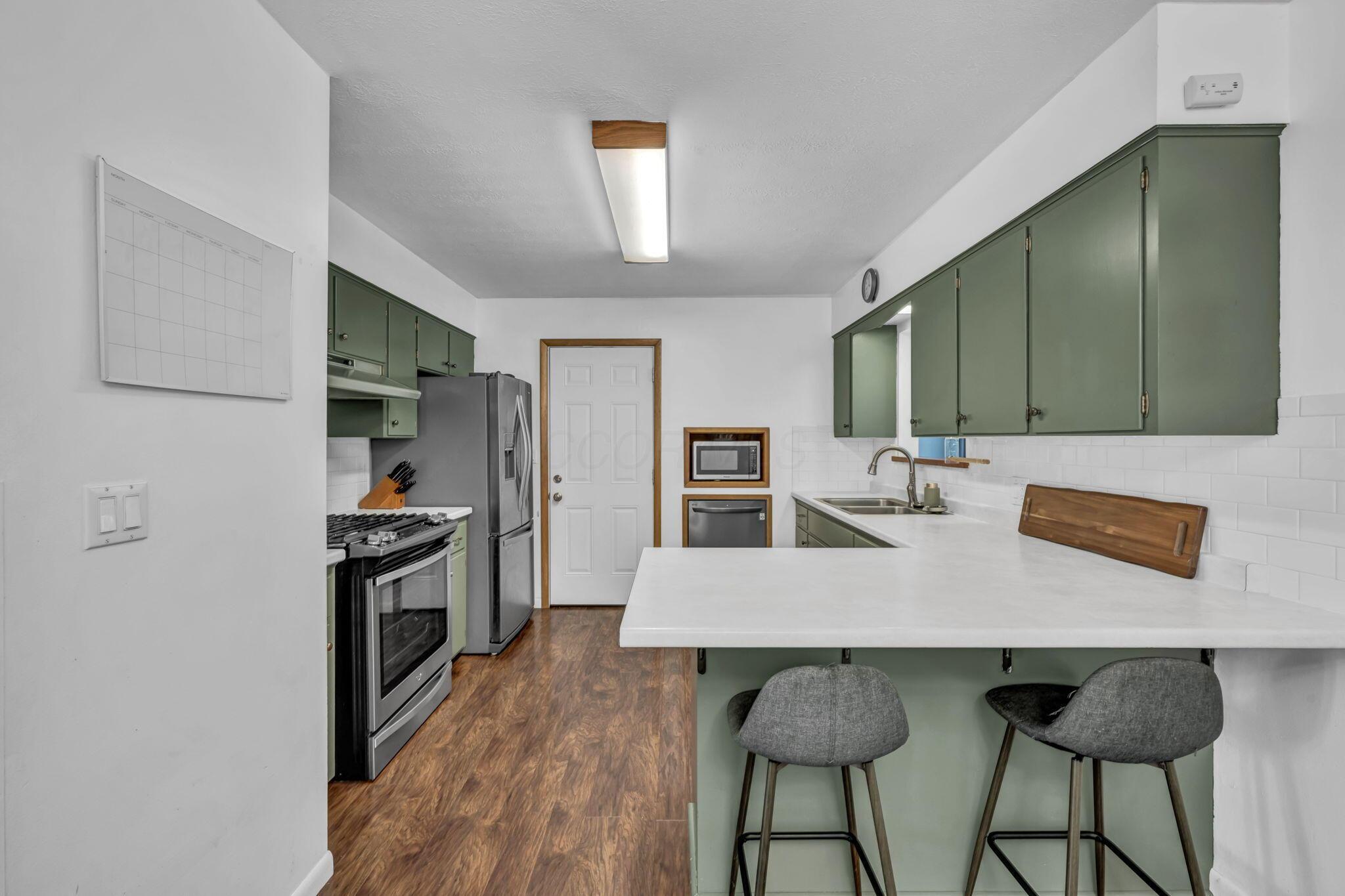 Kitchen with sage-green cabinets, stainless appliances, and a large island with two gray stools.