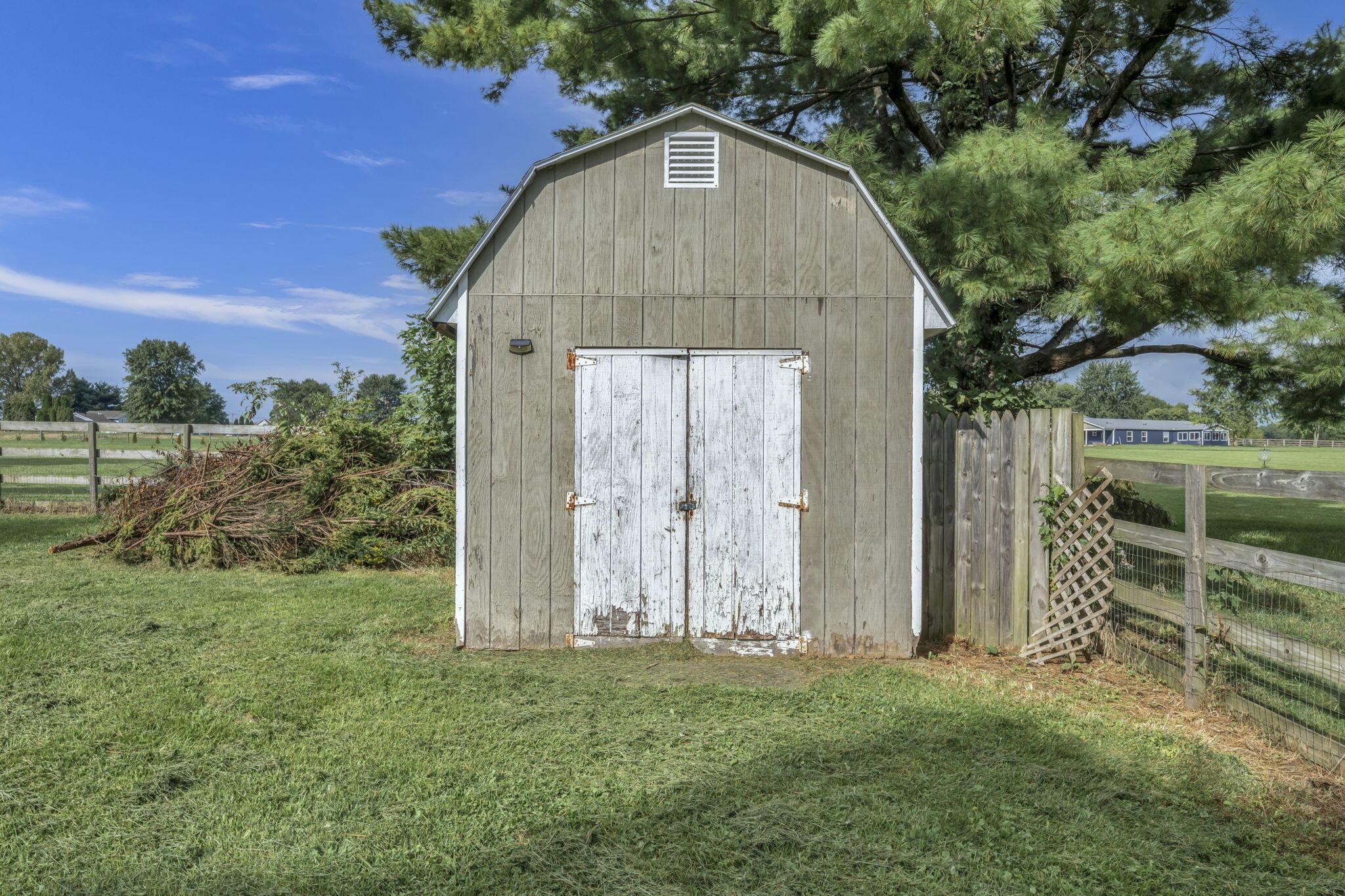 Weathered gray barn with white double doors beside a fenced grassy yard.