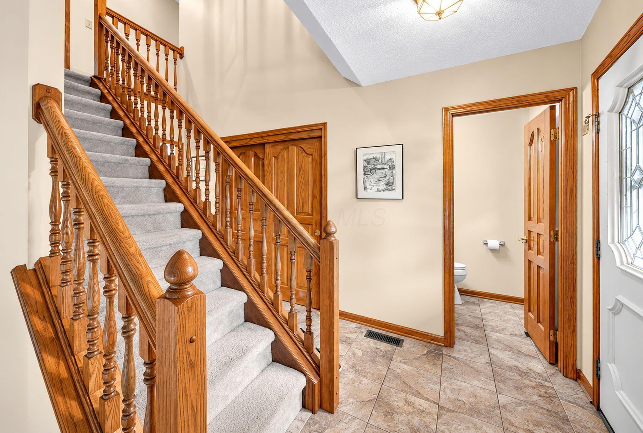 Foyer with a carpeted staircase, ornate wood railings, tiled floor, and doorway to a half bath.