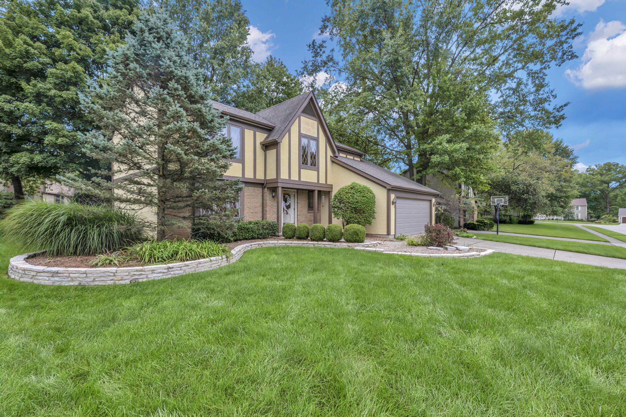Two-story Tudor-style home with an attached garage, mature trees, landscaped beds, and a wide front lawn.