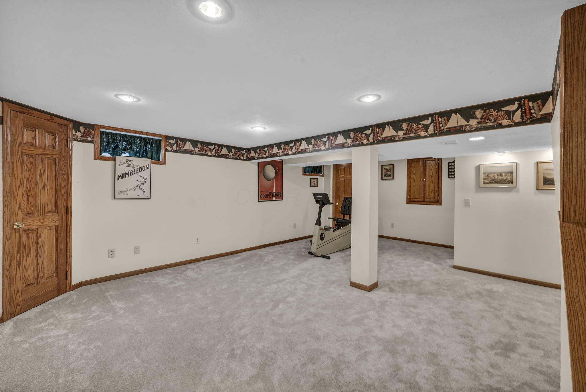Basement recreation room with a stationary bike, gray carpet, wood trim, and sports posters.