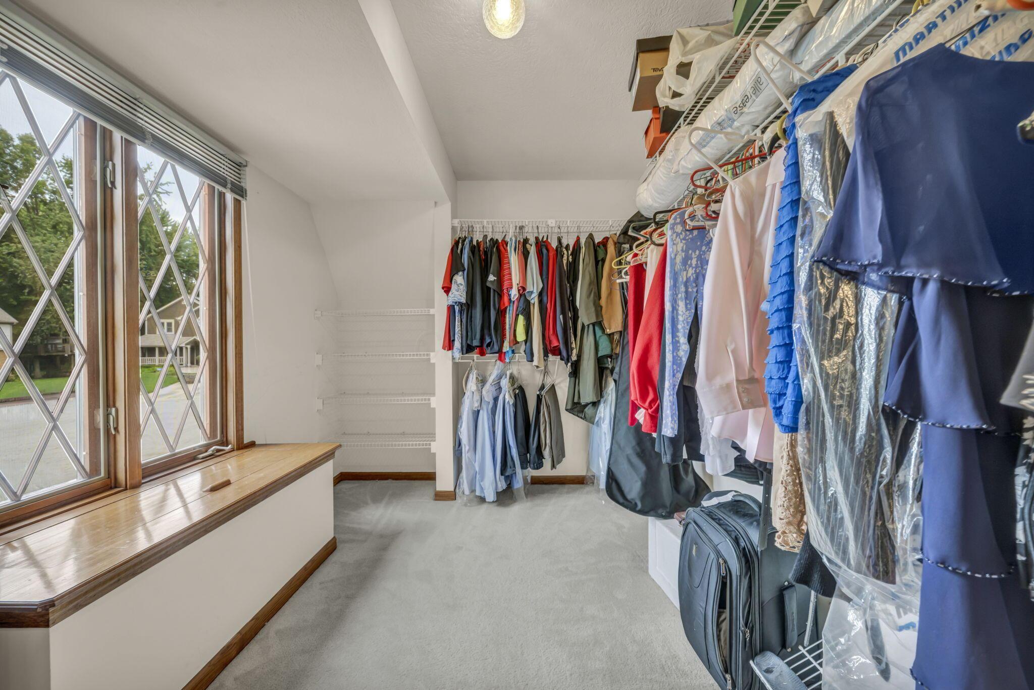Walk-in closet with clothes on racks, luggage, empty shelves, and a large leaded-glass window.