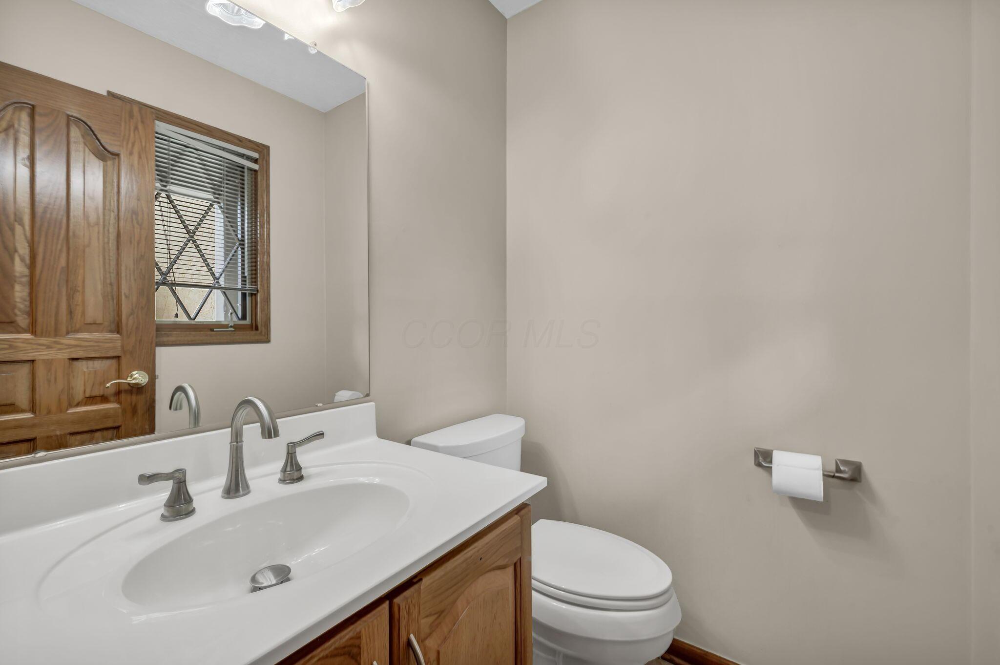 Bathroom with a white vanity, wood cabinetry, large mirror, and window with blinds.