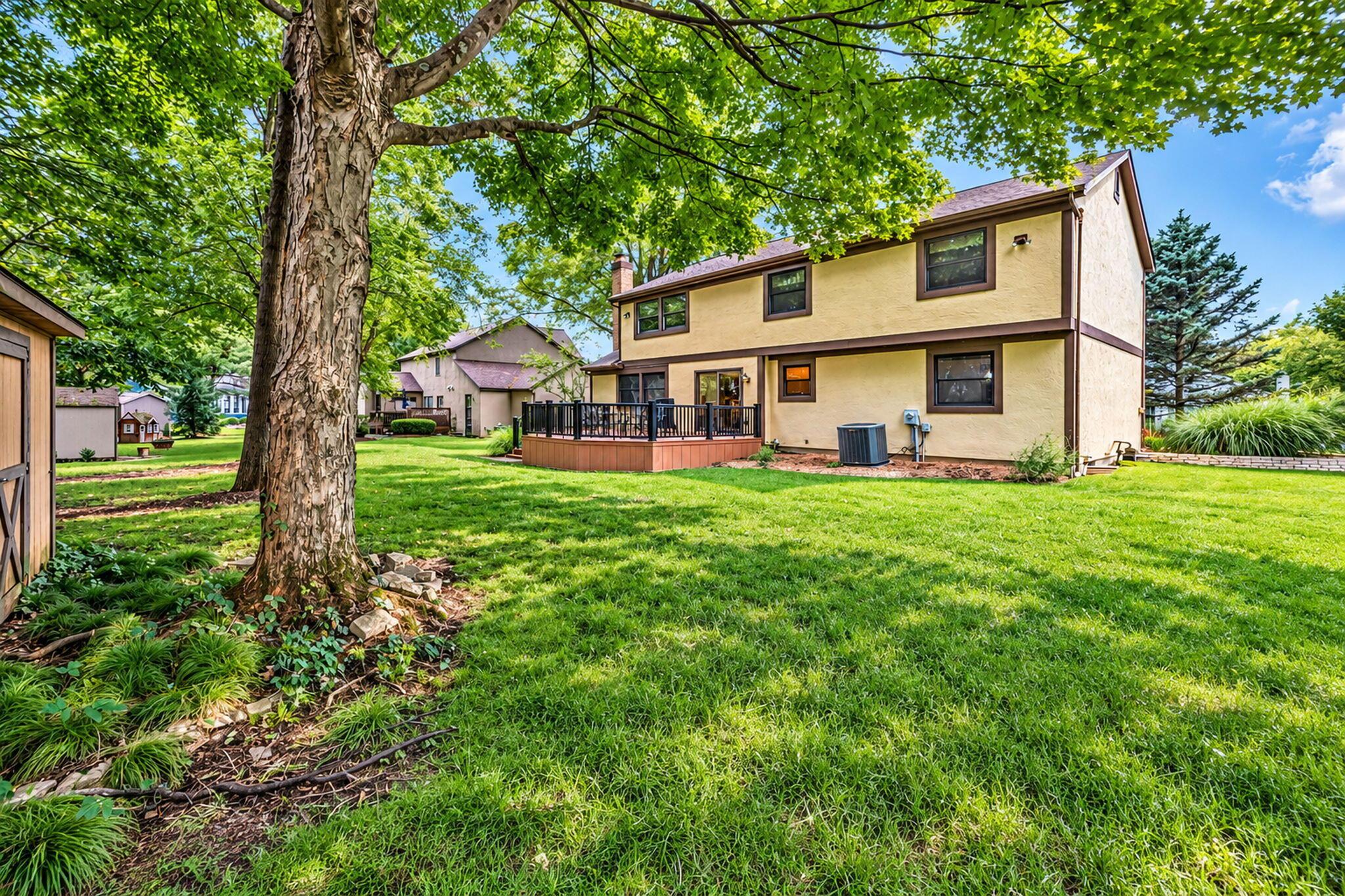 Rear of a two-story tan house with a raised deck and spacious lawn beneath a mature tree.