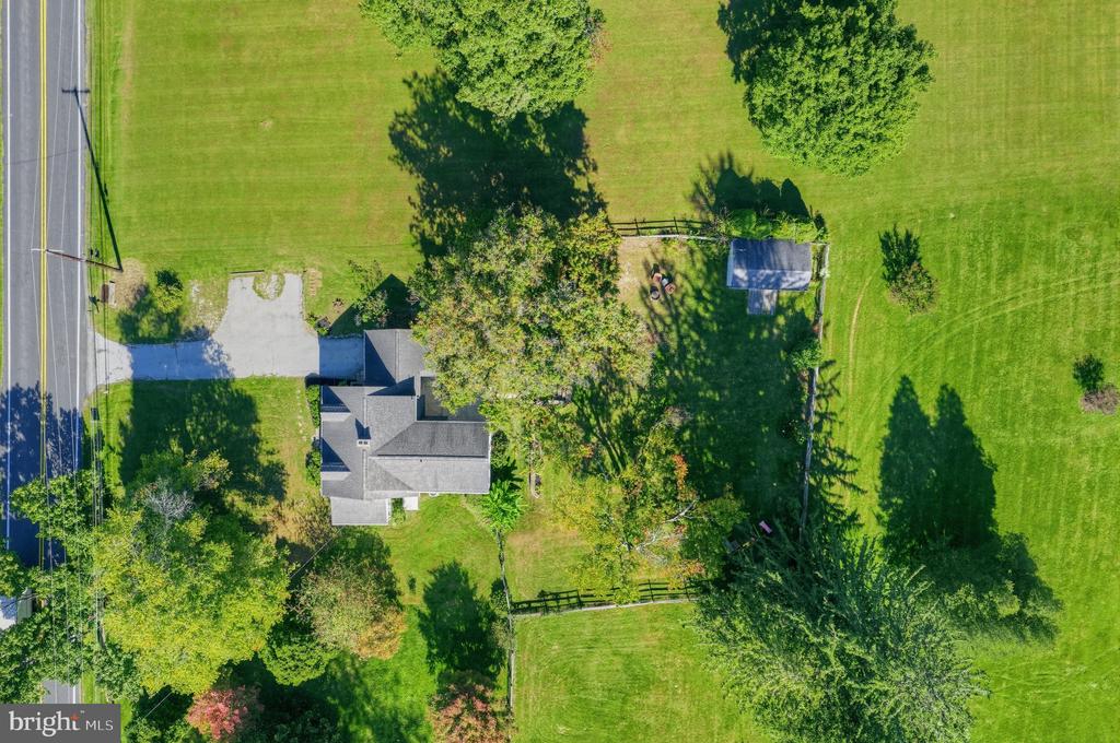 Aerial view of a home surrounded by broad lawns and mature trees, with a driveway and fenced backyard.