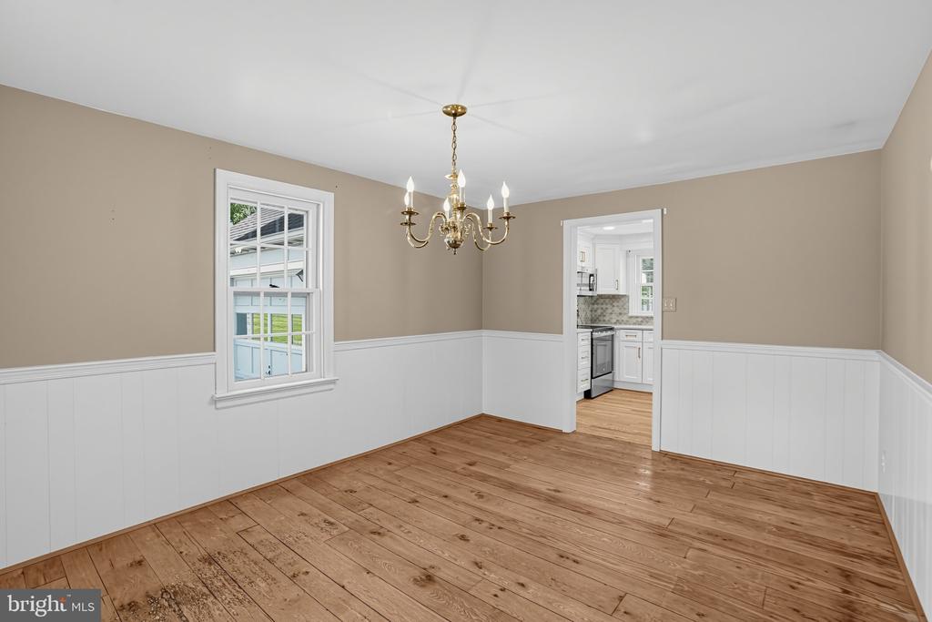 Empty dining room with wood floors, white wainscoting, and a brass chandelier beside the kitchen.