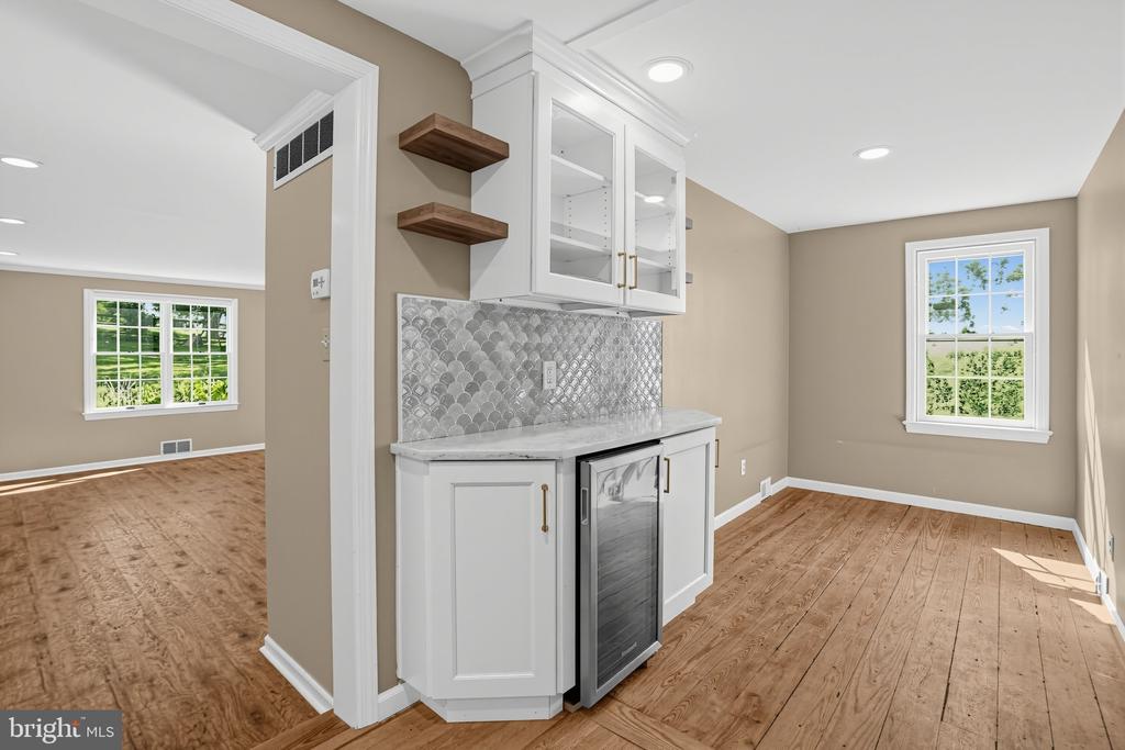 Built-in beverage station with glass-front cabinets and patterned backsplash beside a sunlit hardwood-floored room.