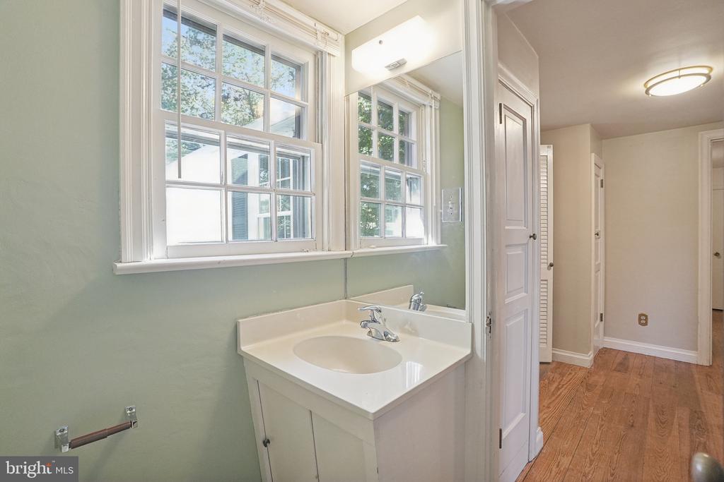 Bathroom vanity beneath a large mirror, beside two tall multi-pane windows and a wood-floored hallway.