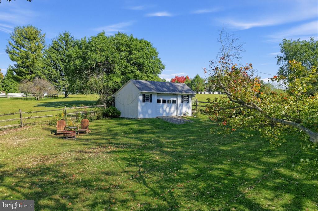 Detached white outbuilding beside a fenced lawn with a fire pit and mature trees.