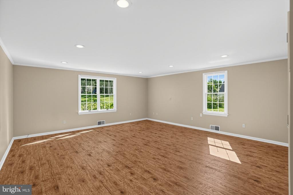 Empty room with brown wood-look flooring, recessed lights, and two windows overlooking greenery.