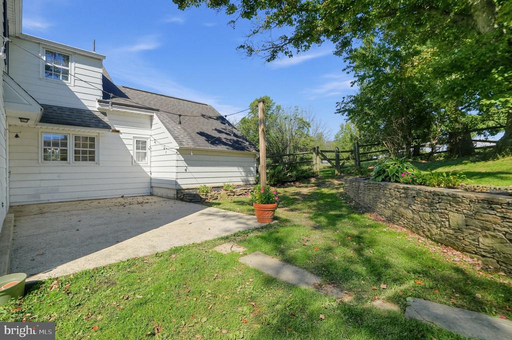 White-sided home beside a concrete patio and grassy yard with a stone retaining wall and mature trees.