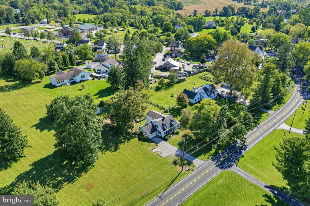 Aerial view of a white house with a dark roof, broad lawn, mature trees, and a curving road nearby.