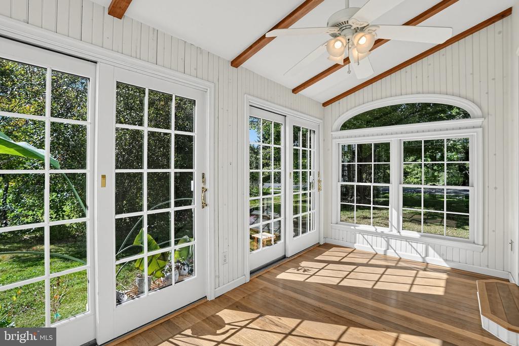 Sunroom with hardwood floors, exposed beams, and large windows and glass doors overlooking a leafy yard.