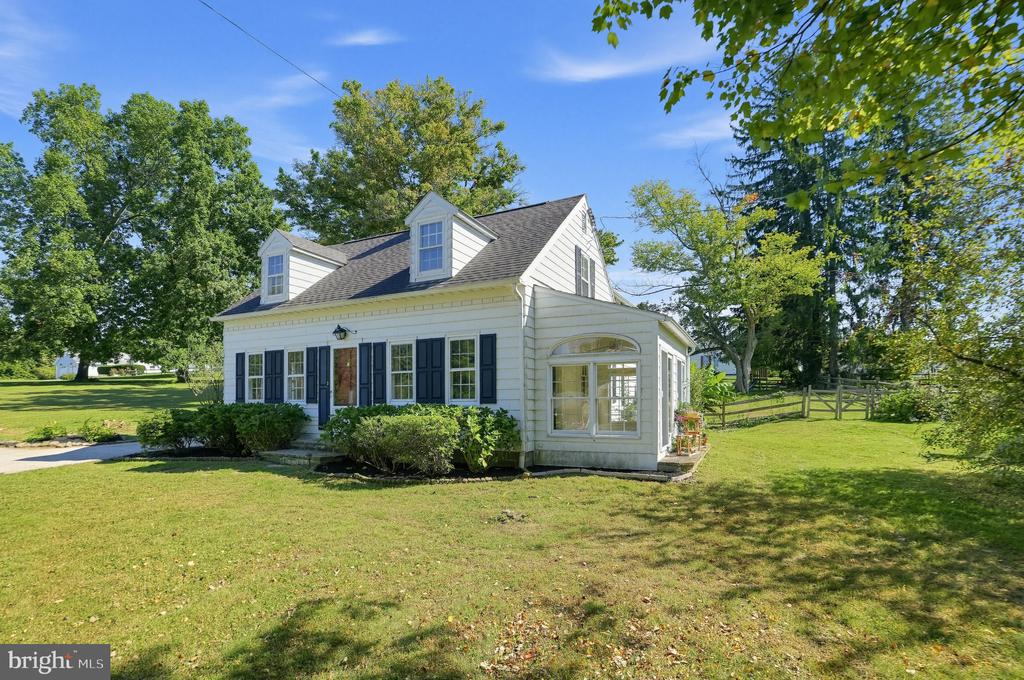White two-story house with dormer windows and dark shutters, set on a broad lawn beneath mature trees.