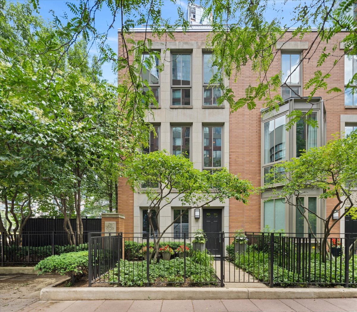 Brick residential building with a fenced, landscaped entrance partly screened by leafy trees.